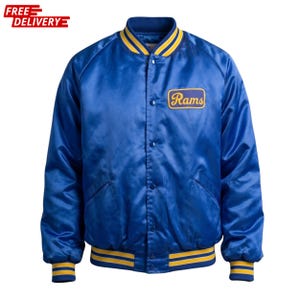 Puede incluir: Chaqueta bomber de satén azul con ribete a rayas amarillas y azules en el cuello, los puños y el dobladillo. La chaqueta presenta un parche que dice "Rams" en letras amarillas. La chaqueta tiene una parte delantera abotonada y dos bolsillos delanteros.