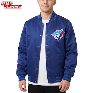 Può includere: Un giubbotto bomber in raso blu reale con chiusura frontale a bottoni automatici, polsini e colletto a coste a righe e una toppa con il logo dei Toronto Blue Jays sul petto sinistro. La giacca è indossata sopra una t-shirt bianca e pantaloni scuri.