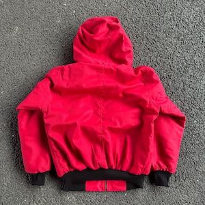 May include: A red hooded jacket with a black bottom hem and black cuffs. The jacket has a zipper closure and is made of a smooth, possibly nylon, material. The hood is up, and the jacket is viewed from the back.