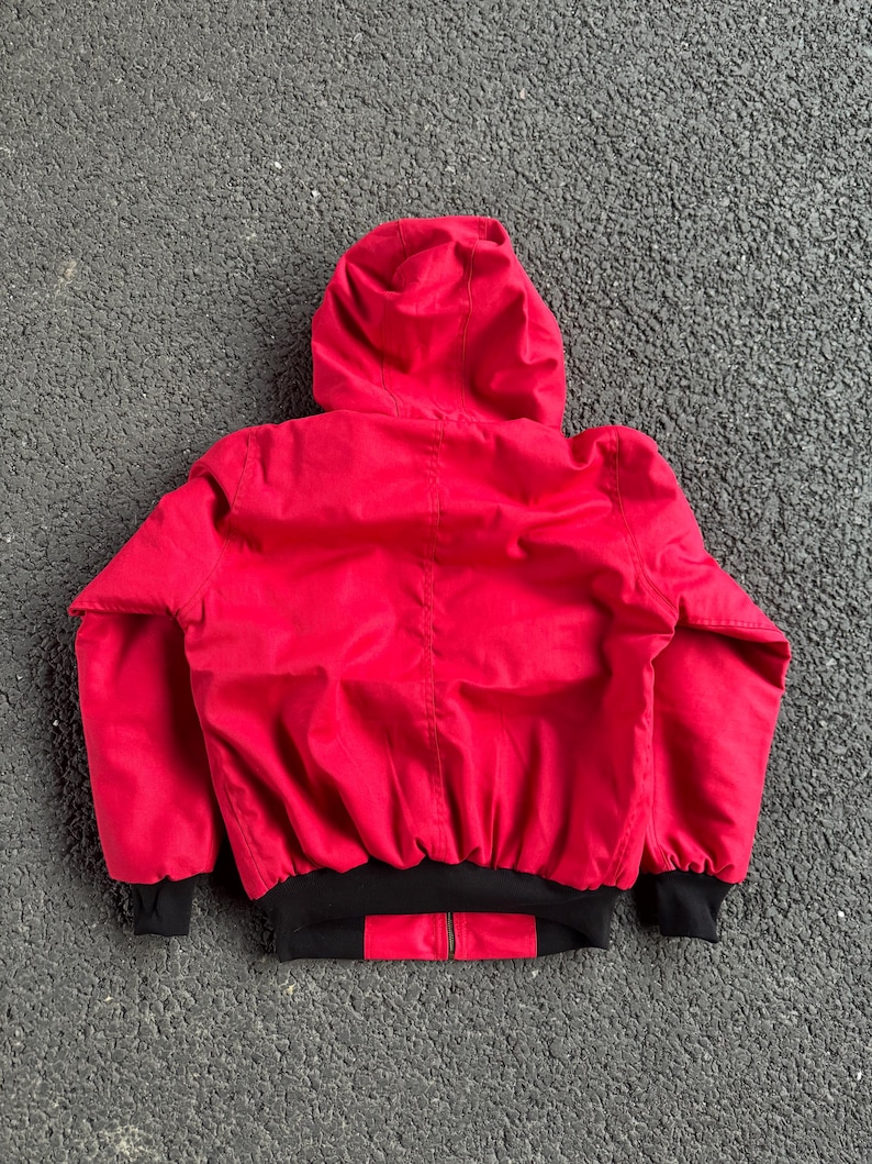 May include: A red hooded jacket with a black bottom hem and black cuffs. The jacket has a zipper closure and is made of a smooth, possibly nylon, material. The hood is up, and the jacket is viewed from the back.