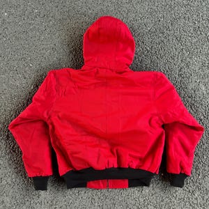 May include: A red hooded jacket with a black bottom hem and cuffs. The jacket is made of a smooth material and has a full zipper closure. The hood is attached to the jacket and is designed to cover the head.