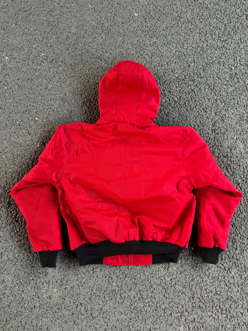 May include: A red hooded jacket with a black bottom hem and cuffs. The jacket is made of a smooth material and has a full zipper closure. The hood is attached to the jacket and is designed to cover the head.