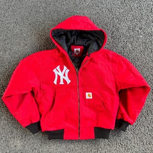 May include: Red hooded jacket with a white New York Yankees logo on the chest. The jacket has a black interior lining, black cuffs, and a full-length zipper. A small Carhartt logo is visible on the front.