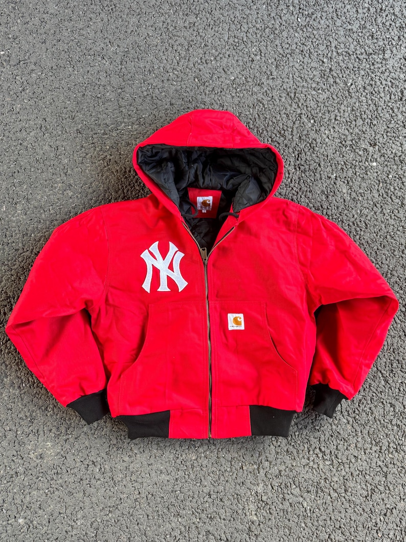 May include: Red hooded jacket with a white New York Yankees logo on the chest. The jacket has a black interior lining, black cuffs, and a full-length zipper. A small Carhartt logo is visible on the front.