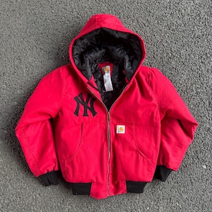 May include: Red hooded jacket with a black lining and a black zipper. The jacket features the New York Yankees logo in black on the chest and a Carhartt logo patch. The jacket has black cuffs and a black bottom hem.
