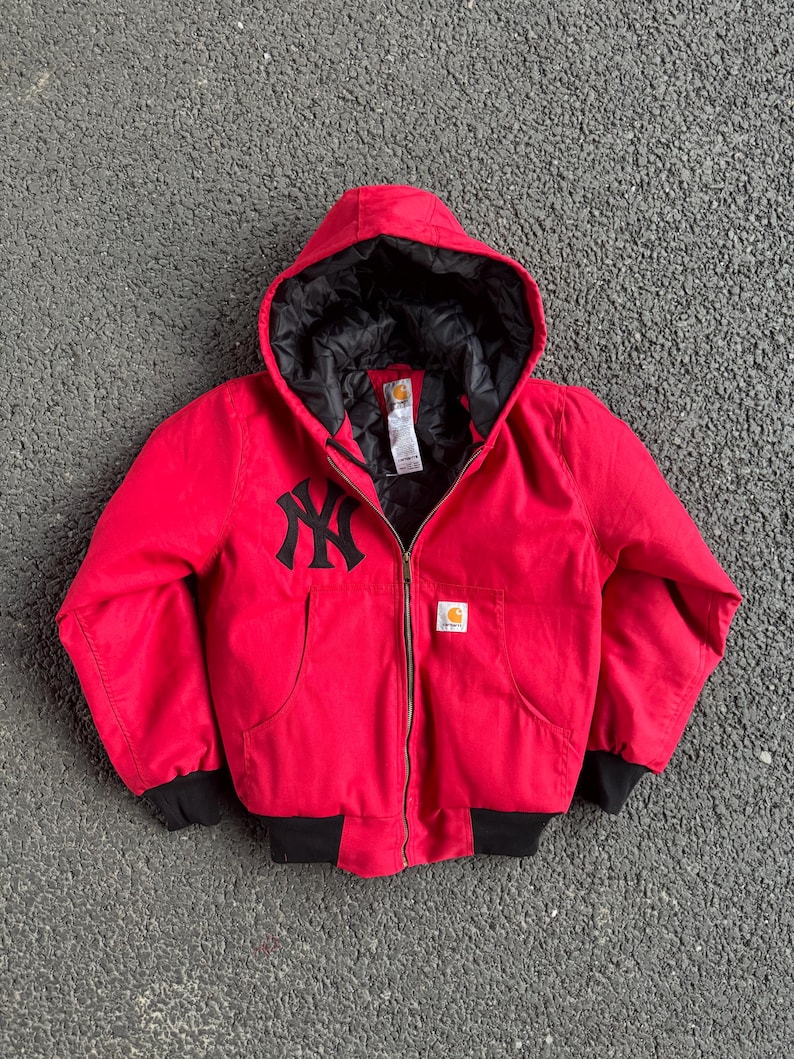 May include: Red hooded jacket with a black lining and a black zipper. The jacket features the New York Yankees logo in black on the chest and a Carhartt logo patch. The jacket has black cuffs and a black bottom hem.