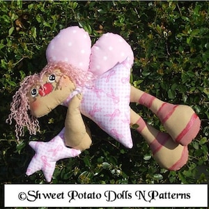 Puede incluir: Una muñeca de tela rosa con alas de lunares rosas y blancos, un vestido de cuadros rosa y blanco con cintas rosas y una estrella rosa con una cinta rosa. La muñeca lleva un vestido de cuadros rosa y blanco con cintas rosas. La muñeca sostiene una estrella rosa con una cinta rosa. La muñeca está sentada en un lecho de hojas verdes.