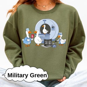 Op de afbeelding: Militairgroen sweatshirt met een cartoonillustratie van eenden in een medische setting. Eén eend houdt een 'L'-bord vast, een andere zit in een CT-scanner en er is een medische röntgenfoto. Het ontwerp bevat ook een eendendokter.