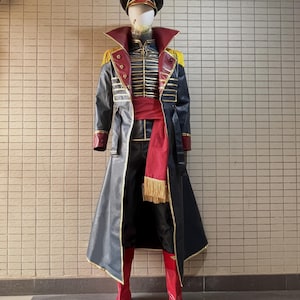 May include: A military-style costume on a mannequin. The outfit includes a black hat, a long black coat with gold trim, a red sash, and red boots. The coat has a high collar and gold epaulets. The mannequin is standing against a tiled wall.