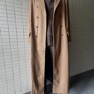 Op de afbeelding: Een lange, beige, dubbelrijige overjas gedragen over een bruin vest met krijtstreep en bijpassende broek. Het ensemble bevat een wit overhemd met kraag en een donkere das met patroon, getoond op een etalagepop voor een betegelde muur.