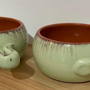 Puede incluir: Dos cuencos de cerámica para sopa con asas. Los cuencos son de color verde claro con interior marrón y borde marrón. Las asas tienen un diseño único. Los cuencos están sobre una superficie de madera.