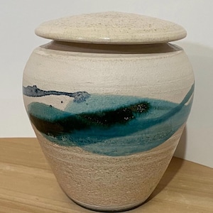 Può includere: Un'urna in ceramica con coperchio. L'urna è bianco sporco con una superficie strutturata e una forma arrotondata. Presenta un disegno dipinto in turchese e blu scuro al centro. Il coperchio è anch'esso bianco sporco e strutturato.