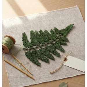 May include: A handmade, green crochet fern leaf rests on a neutral linen surface. Wooden crochet hooks and a spool of green thread are nearby. A blank tag with string is also present, suggesting a handcrafted item.