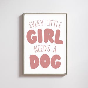 Op de afbeelding: Een ingelijste print met de tekst "EVERY LITTLE GIRL NEEDS A DOG" in een speelse, roze lettertype. De woorden staan gecentreerd op een witte achtergrond, omlijst door een grijze lijst. Een charmant stukje wandkunst voor een kinderkamer.