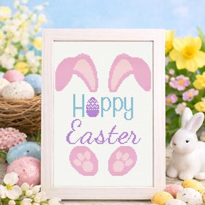 May include: A framed cross-stitch design with the words "Hoppy Easter" in blue and purple, featuring bunny ears and paws. The frame is white, and the background includes Easter eggs, flowers, and a ceramic bunny.