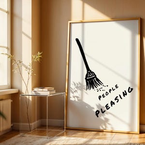 May include: A framed art print featuring a black illustration of a broom with the words "PEOPLE PLEASING" in a handwritten font. The artwork is set against a white background with a gold frame, leaning against a beige wall.