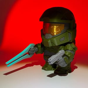 May include: A green and gray figurine of a character from the Halo video game series. The figure is holding a blue energy sword in one hand and a gray pistol in the other. The background is a gradient of red.