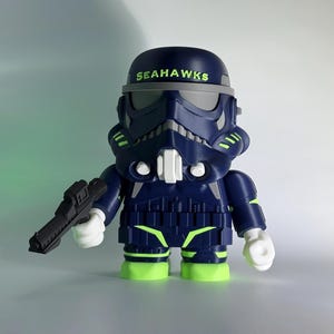 May include: A navy blue and lime green collectible figurine resembling a stormtrooper helmet and armor. The helmet features the word SEAHAWKS in lime green text across the forehead. The figure holds a black blaster in its right hand.