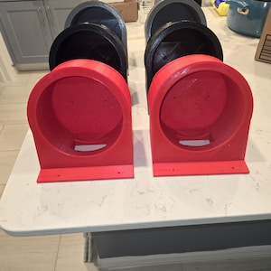 May include: A collection of 3D-printed, semi-circular objects in red and black. The red objects are in the foreground, with a circular opening and a rectangular base. The black objects are in the background, with a similar design. The objects are arranged on a white surface.