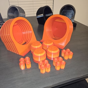 Puede incluir: Un surtido de objetos naranjas y amarillos impresos en 3D. La colección incluye estructuras grandes, en capas, en forma de cono, piezas cilíndricas con una 'D' en la parte superior y piezas hexagonales más pequeñas. Varios objetos negros y grises están en el fondo.