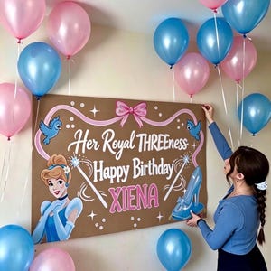 May include: A rectangular brown sign featuring a Cinderella illustration, a glass slipper, blue birds, and a pink bow. The text reads Her Royal THREENESS Happy Birthday XIENA. The sign is surrounded by pink and blue balloons on white strings.
