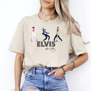 May include: Beige t-shirt featuring three stylized illustrations of Elvis Presley in different performance outfits. The word "ELVIS" is printed in a dotted font, with Elvis's signature below. The shirt is worn by a person in blue jeans.