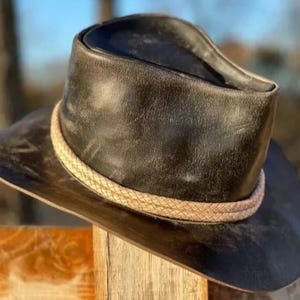 Op de afbeelding: Een zwarte leren hoed met een gevlochten bruin bandje. De hoed heeft een brede rand en een afgeronde kroon. De hoed ligt op een houten oppervlak. De hoed lijkt nieuw te zijn.