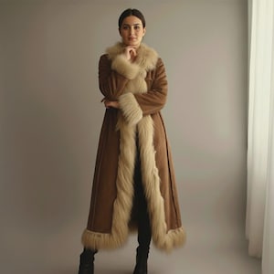 Może przedstawiać: Długi brązowy płaszcz z kremowym futrzanym wykończeniem wokół kołnierza, mankietów i przodu. Płaszcz sięga do kolan i ma klasyczny, elegancki krój. Kobieta na zdjęciu ma na sobie czarne buty.