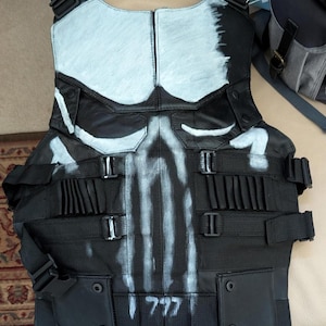 Könnte beinhalten: Schwarze taktische Weste mit weißem Totenkopf-Design auf der Vorderseite. Die Weste hat verstellbare Riemen, Taschen und die Zahl "777" auf der Unterseite. Die Oberseite der Weste ist hellblau lackiert.