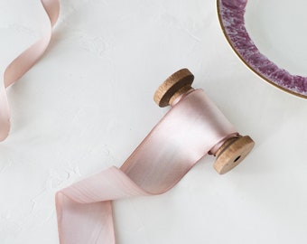 Puede incluir: Un carrete de cinta rosa pálido con extremos de madera, parcialmente desenrollado, se encuentra sobre una superficie blanca. También hay un rollo de cinta a juego y un plato decorativo con borde morado y dorado, lo que sugiere un tema artesanal o decorativo.