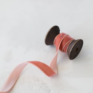 May include: A spool of dusty rose velvet ribbon with a pin on the spool. The ribbon is unraveled and lying on a white surface.