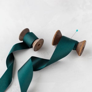 May include: Two spools of teal green ribbon with a pin on one spool. The ribbon is unraveled and laying on a white surface.