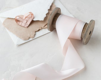 Könnte beinhalten: Eine Spule mit blassrosa Satinband mit einer Holzspule. Das Band ist abgewickelt und liegt auf einer weißen Oberfläche. Ein kleines, gefaltetes, herzförmiges Stück Papier befindet sich im Hintergrund.