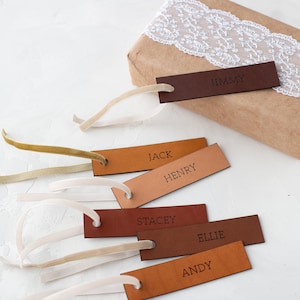May include: A collection of rectangular leather gift tags in various shades of brown, each featuring a name embossed in capital letters: Jimmy, Jack, Henry, Stacey, Ellie, and Andy. Each tag includes a thin velvet ribbon loop for attachment.