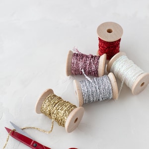 Puede incluir: Cinco carretes de madera envueltos con cordón metálico brillante en rojo, rosa oscuro, plata, oro y blanco. Unas tijeras rojas descansan cerca del cordón dorado, con el hilo dorado extendiéndose desde el carrete a través de las hojas abiertas.