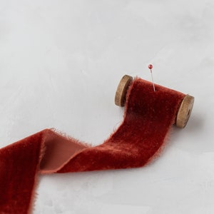 Puede incluir: Un carrete de cinta de terciopelo color óxido con un borde deshilachado y un alfiler con cabeza roja. La cinta está parcialmente desenrollada, revelando su textura. El carrete de madera es visible, añadiendo un toque rústico. Ideal para manualidades y proyectos decorativos.