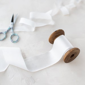 May include: A spool of white satin ribbon with a wooden spool and a pair of scissors on a white textured surface.