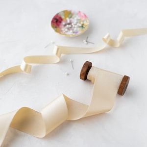 May include: A spool of ivory satin ribbon with a wooden spool, a small dish with white pins, and a few loose pins on a white surface.