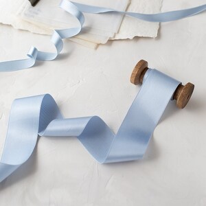 Puede incluir: Un carrete de cinta de satén azul claro con un carrete de madera. La cinta está enrollada alrededor del carrete y está lista para ser utilizada para manualidades o decoración.