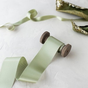 May include: A spool of pale green satin ribbon with wooden ends, partially unrolled. A length of the ribbon is unraveled, and a piece of olive green fabric with gold embellishments is in the background. The ribbon is on a white surface.