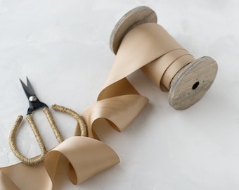 May include: A spool of beige satin ribbon with a wooden spool and a pair of gold-trimmed scissors.