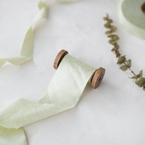 May include: A spool of pale green silk ribbon with wooden ends. The ribbon is unraveled and draped on a white surface.