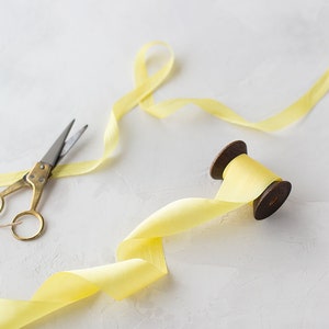 May include: A spool of yellow ribbon with a wooden spool and a pair of gold scissors on a white surface.