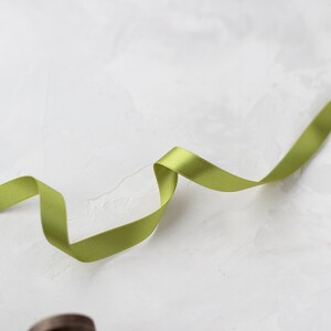 May include: A spool of green satin ribbon with a small amount of ribbon unraveled and laying on a white textured surface.