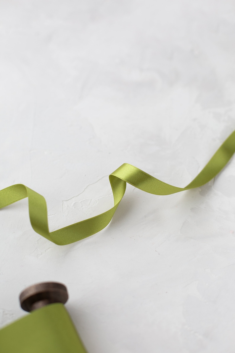 May include: A spool of green satin ribbon with a small amount of ribbon unraveled and laying on a white textured surface.