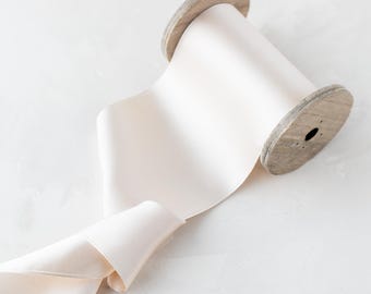 May include: A spool of off-white satin ribbon on a wooden spool. The ribbon is unfurled and draped over the spool.