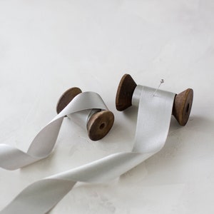 Könnte beinhalten: Zwei Spulen mit weißem Satinband, eine mit einer Stecknadel, auf einer weißen Oberfläche.