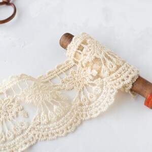 May include: A spool of off-white lace trim with a delicate floral pattern. The lace is wound around a wooden spool with a red accent.