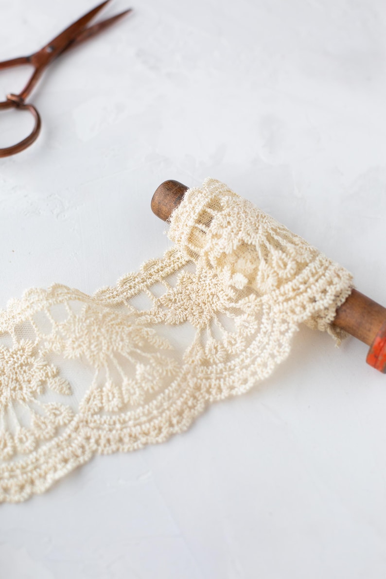 May include: A spool of off-white lace trim with a delicate floral pattern. The lace is wound around a wooden spool with a red accent.