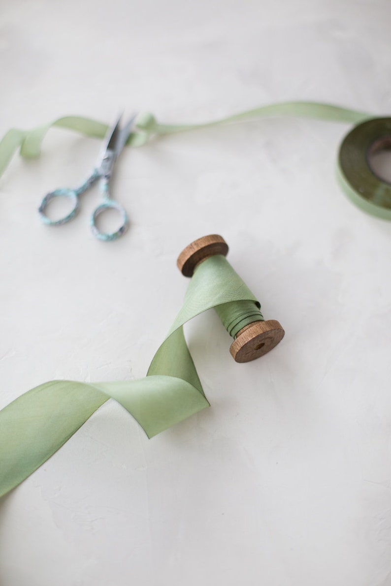 May include: A spool of sage green ribbon with a wooden spool and a pair of silver scissors.